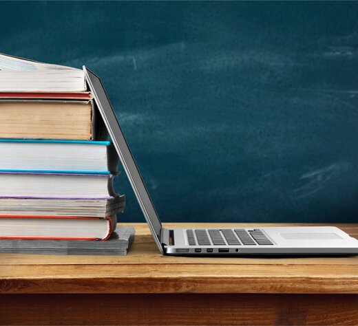 Auf dem Bild ist ein Bücherstapel zu sehen. Ein Laptop lehnt am Bücherstapel, im Hintergrund ist eine Schultafel.
