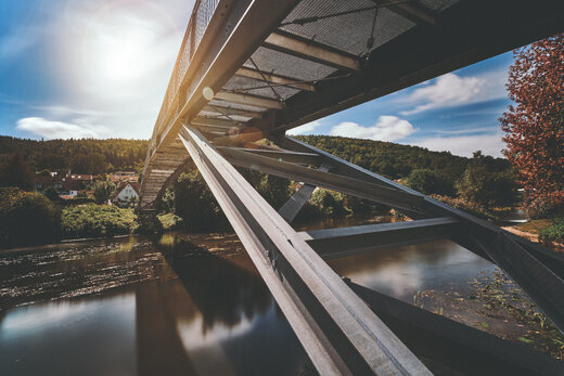 Moderne Stahlbrücke über einem Fluss, aufgenommen von der Unterseite. Im Hintergrund sind eine bewaldete Landschaft und einige Häuser bei sonnigem Wetter zu sehen.