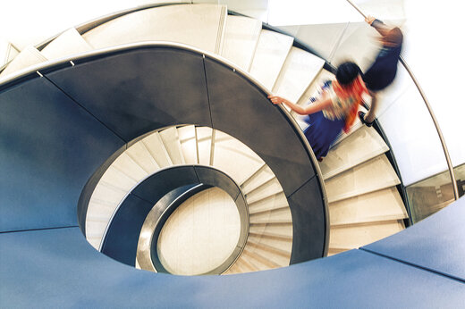 Draufsicht auf eine moderne, geschwungene Wendeltreppe. Zwei Personen bewegen sich unscharf die Treppe hinunter, wodurch Dynamik und Bewegung vermittelt werden.