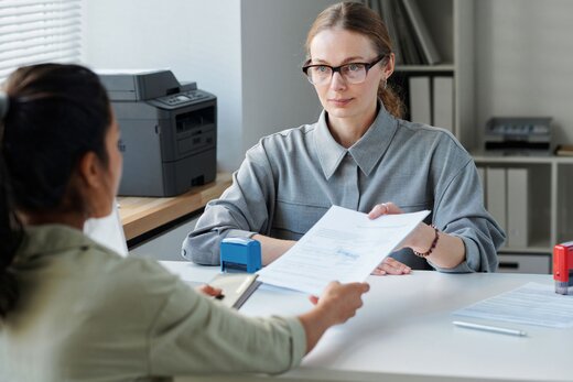 Das Bild zeigt zwei Frauen in einer Behörde: Die eine Frau, die Behördenmitarbeiterin, übergibt Dokumente an die andere Frau, die Antragstellerin.