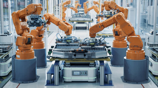 Orangefarbene Roboterarme arbeiten an einer automatisierten Produktionslinie in einer hellen Fabrikhalle. Sie greifen und montieren Bauteile auf Fertigungsstationen, während weitere Roboter im Hintergrund parallel arbeiten.